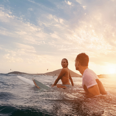 A woman smiles at a man while sitting on surf boards in the sunset.