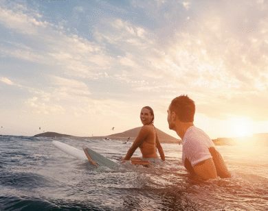 A woman smiles at a man while sitting on surf boards in the sunset.