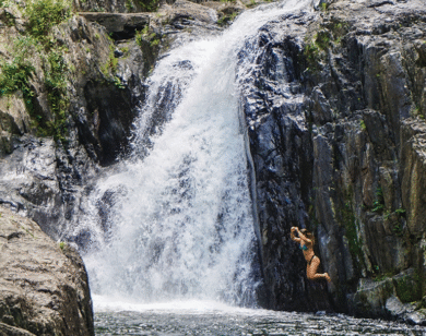 Young woman jumps from a rock into the water below a waterfall.